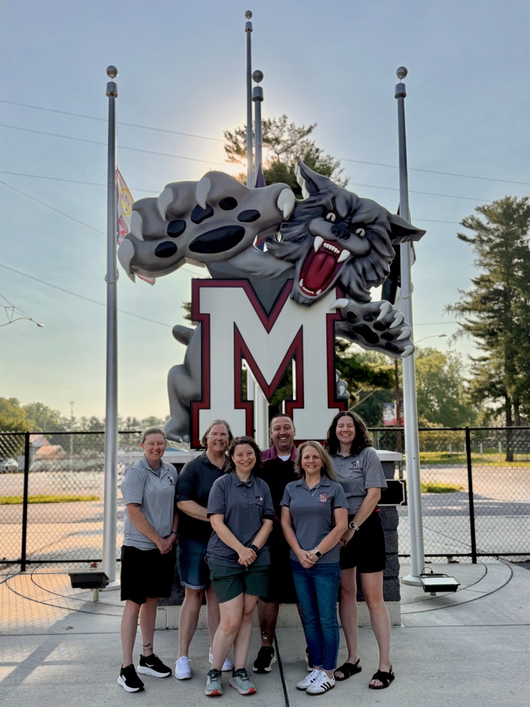 A group of adults wearing gray and maroon shirts, standing in front of a large wildcat M sign.