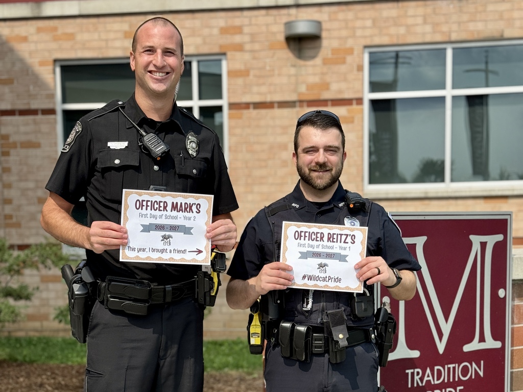 Two police officers hold First day of school signs in front of a school building.