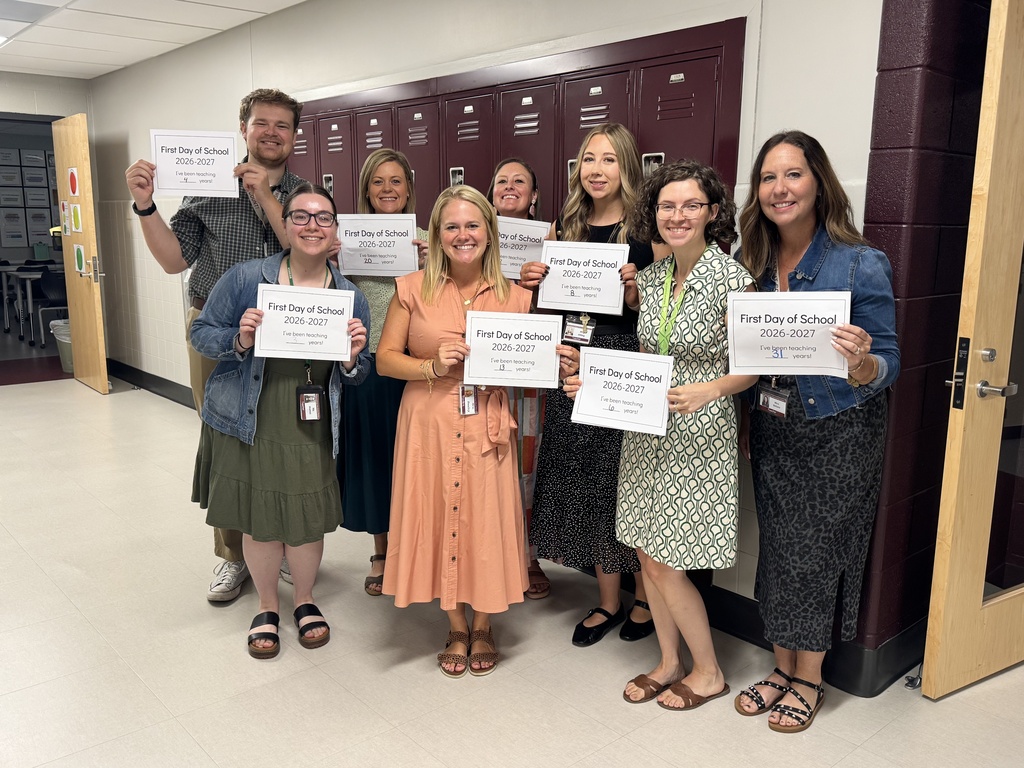a group of teachers stand in front of lockers and hold first day of school signs.