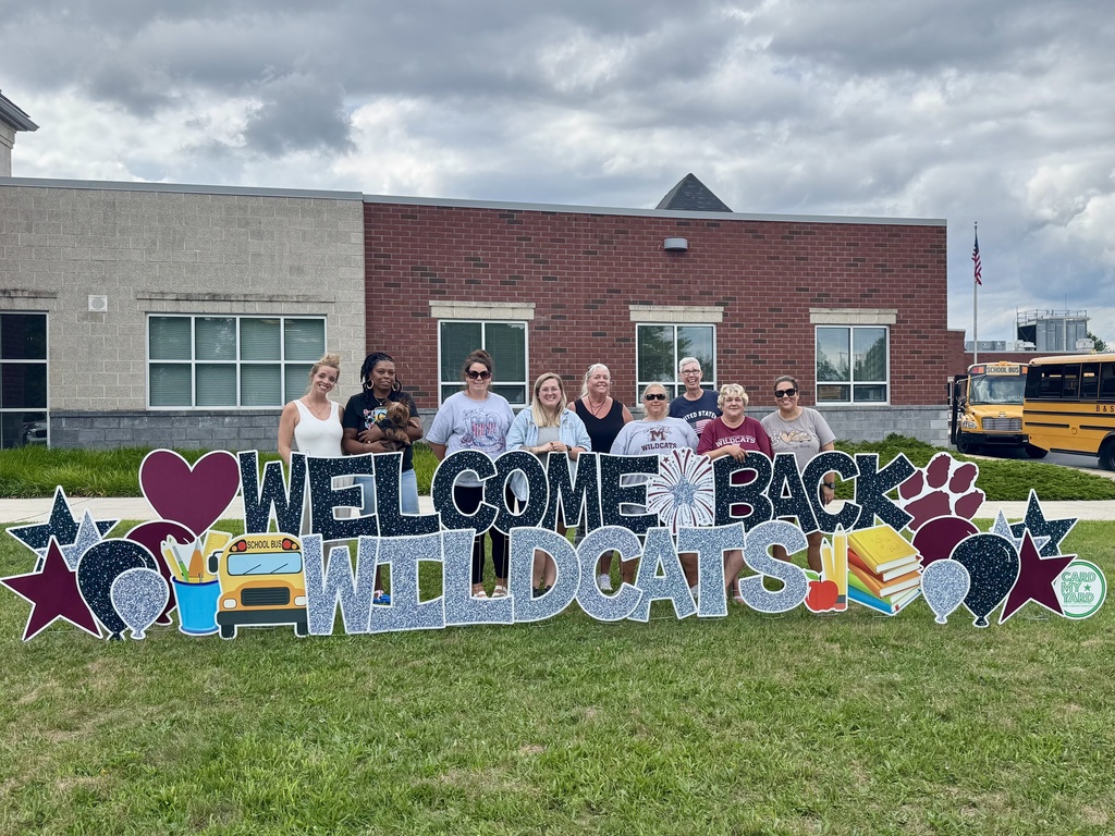 A group of bus drivers standing behind a Welcome Back Wildcats yard sign.