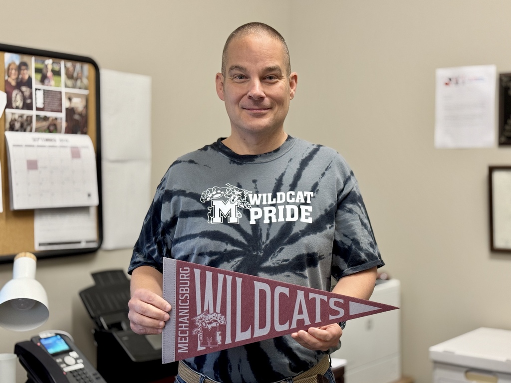 A man proudly wears a steel colored Wildcat shirt while holding a matching maroon pennant.