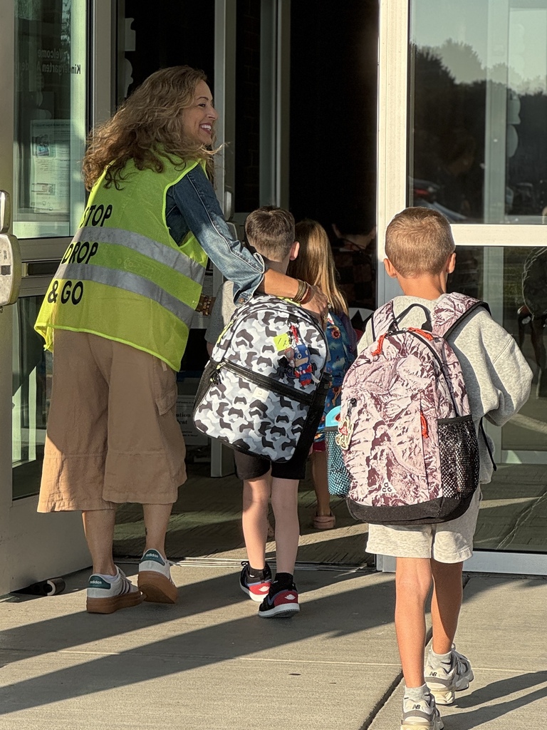 A staff member wearing a bright yellow vest greets students at the start of a school day.