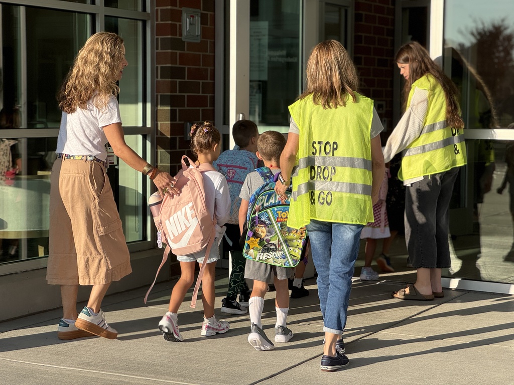 A group of staff members in bright yellow Stop Drop & Go vests help students into a school.