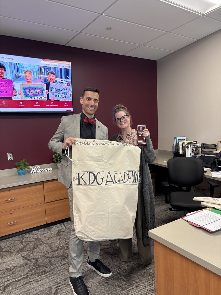 Two adults stand in an office, one holds a hand decorated mail bag and the other a pair of socks.