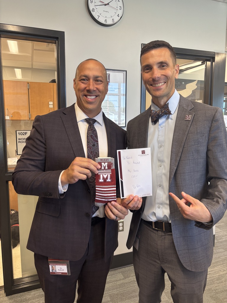 Two men in suits stand side by side, one holding a pair of socks and the other a handwritten note.