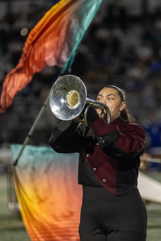 A member of a high school marching band performs while colorful flags are on display behind her.