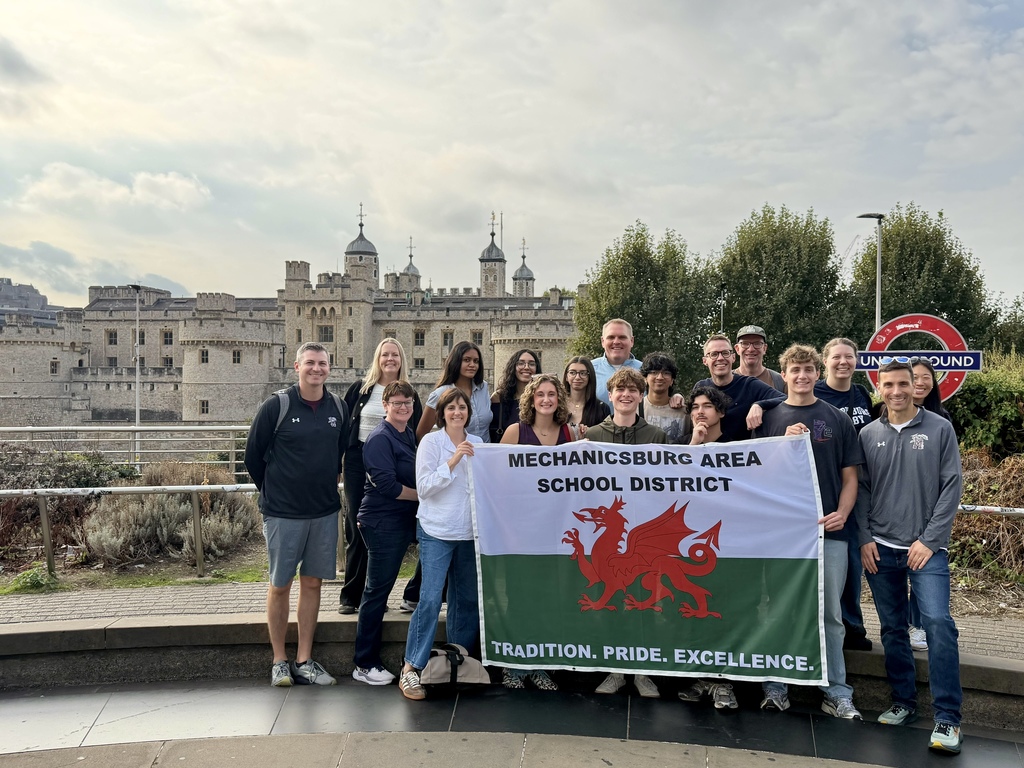 A group of students and educators stand in front of the Tower of London while holding a flag that says Mechanicsburg Area School District.