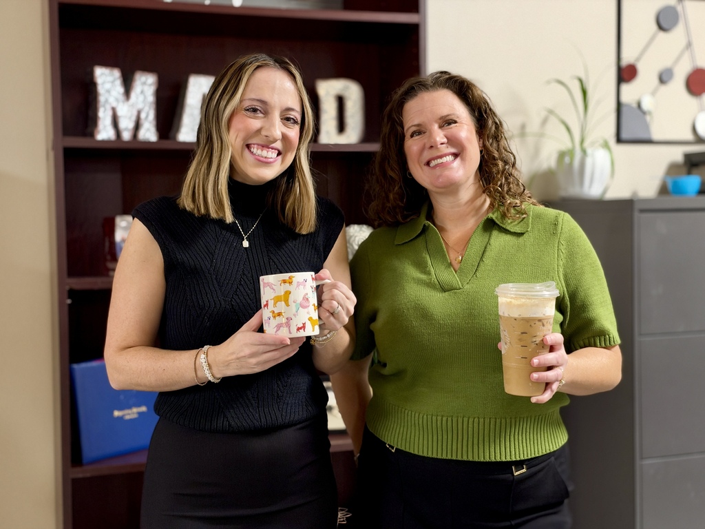 Two ladies stand in front of a bookcase while holding cups of coffee.