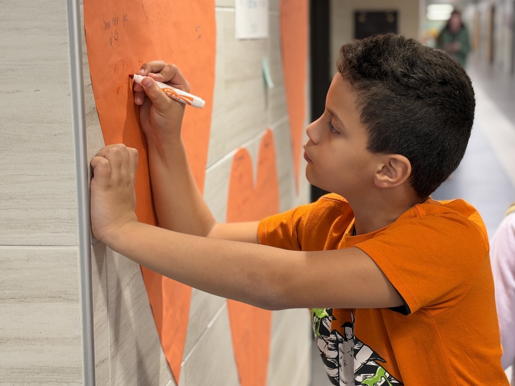Students write kind words and notes of encouragement on an orange heart taped to a school wall.