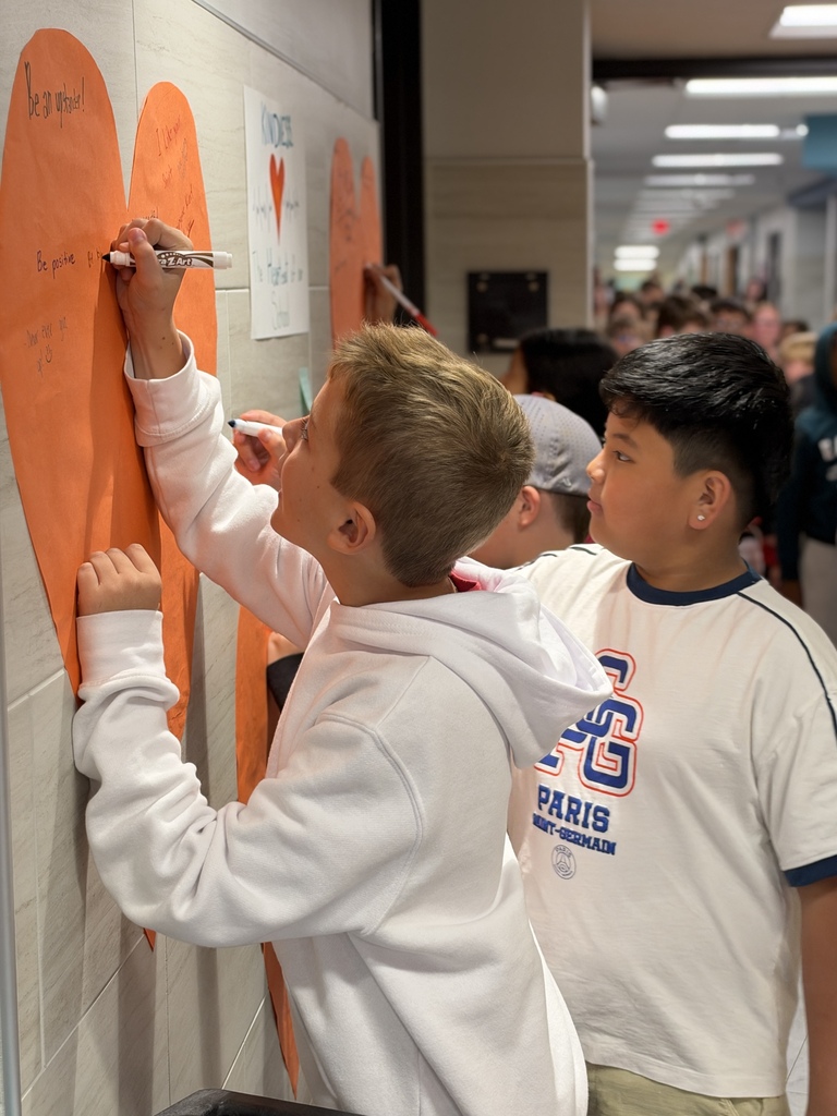 Students write kind words and notes of encouragement on an orange heart taped to a school wall.