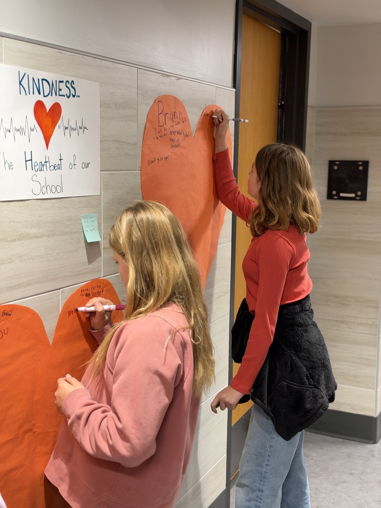 Students write kind words and notes of encouragement on an orange heart taped to a school wall.