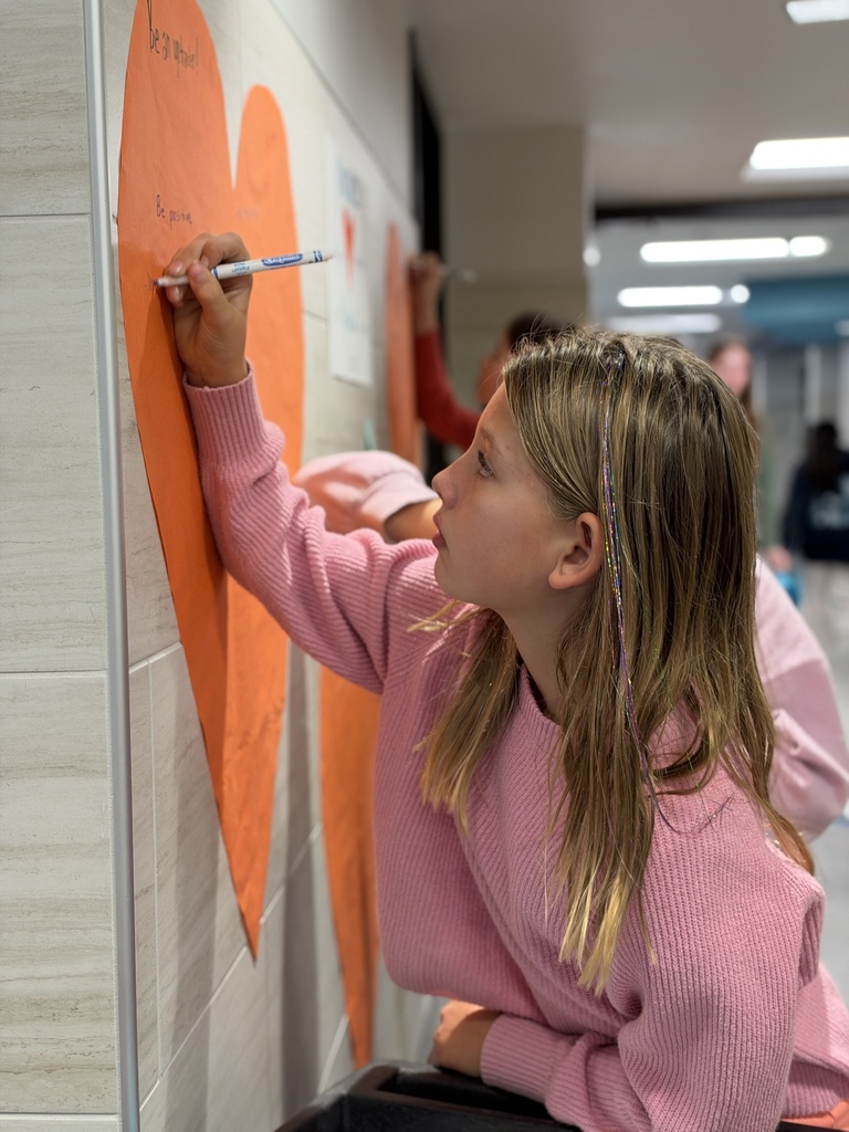 Students write kind words and notes of encouragement on an orange heart taped to a school wall.