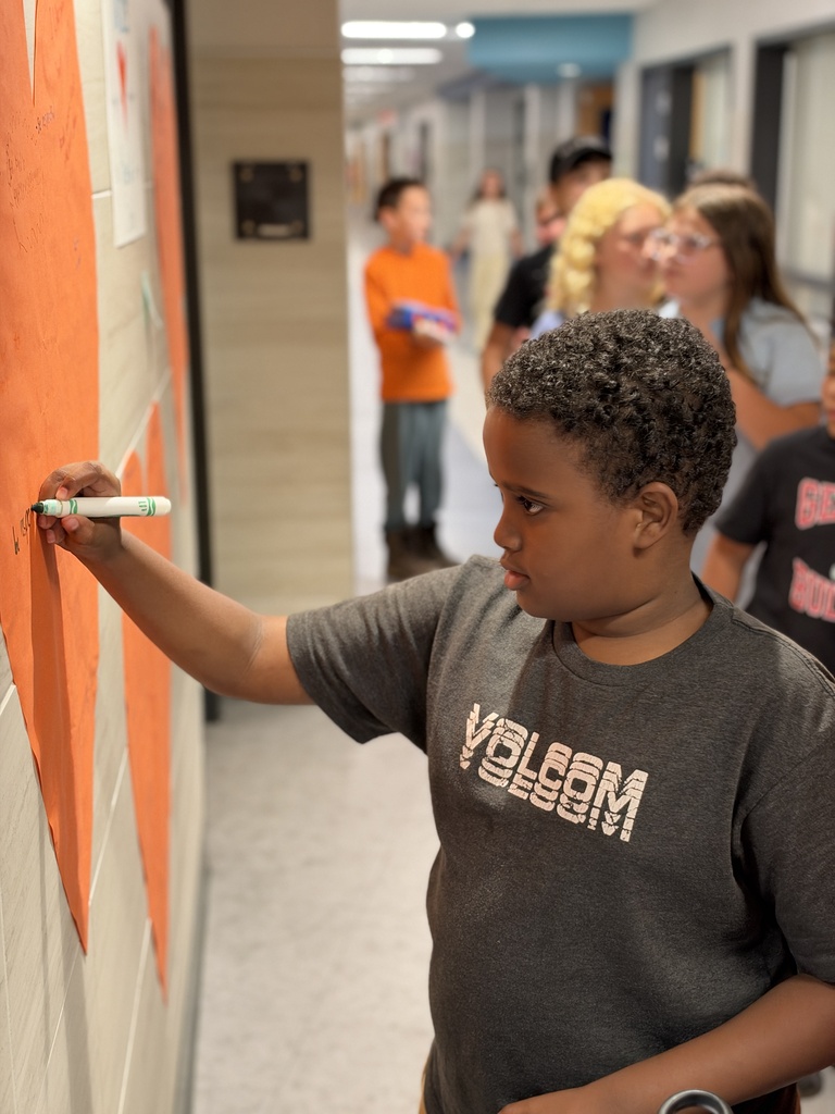 Students write kind words and notes of encouragement on an orange heart taped to a school wall.