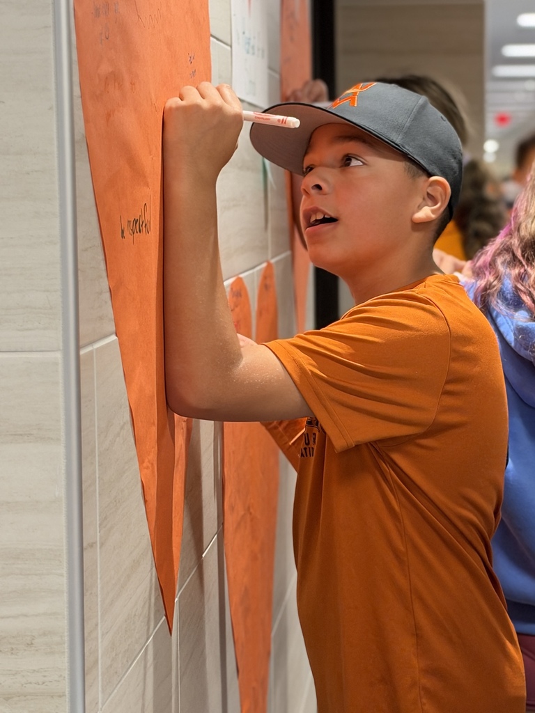 Students write kind words and notes of encouragement on an orange heart taped to a school wall.