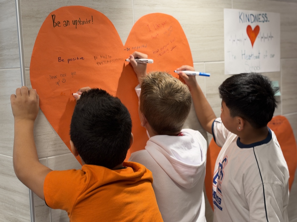 Students write kind words and notes of encouragement on an orange heart taped to a school wall.