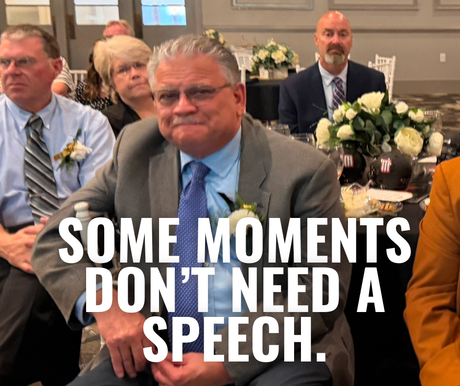 Several people sit in a room with a table, a flower arrangement, and a quote reading "Some moments don't need a speech."