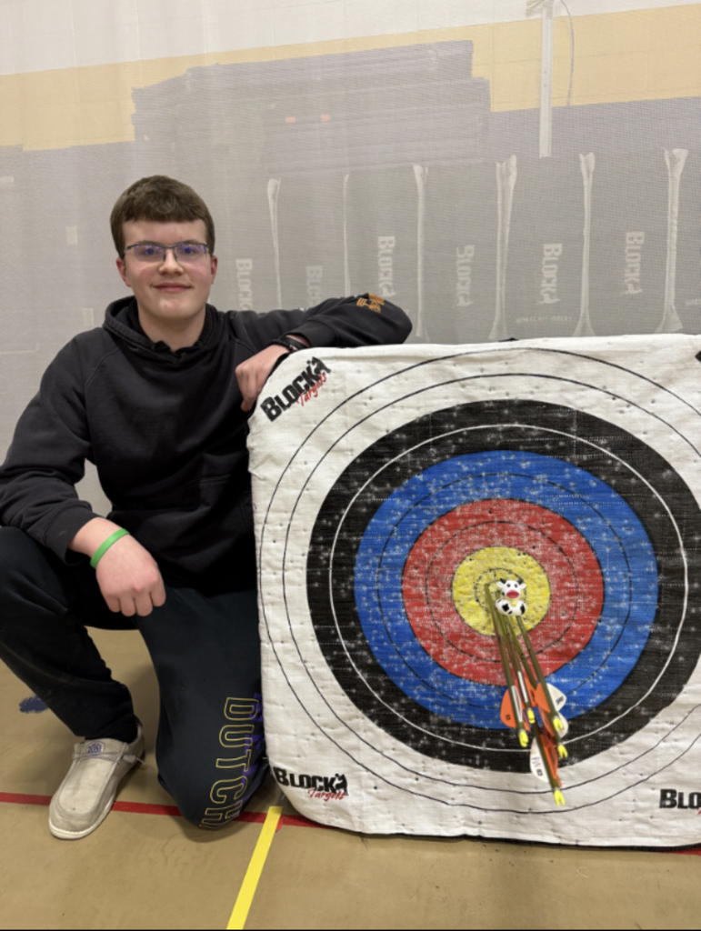 boy with black sweats posed by an archery target with a perfect score and a cow duck on the arrows