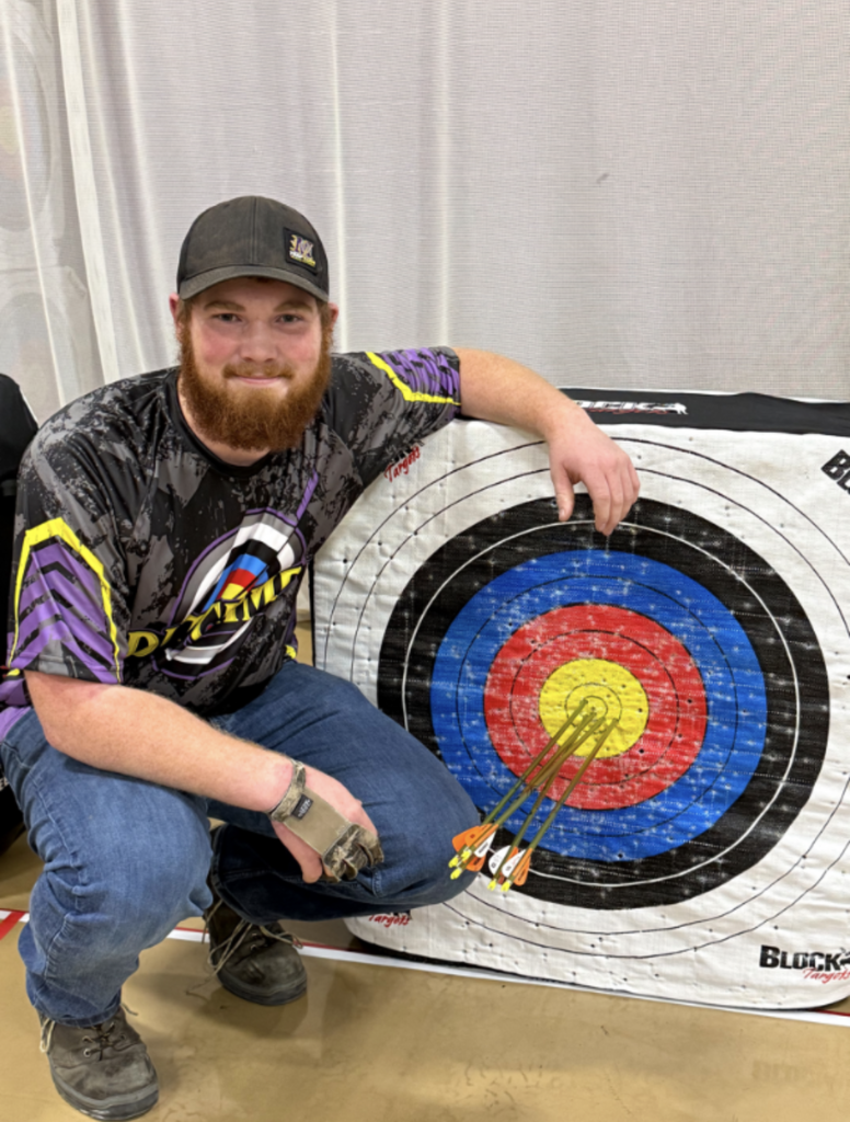 High School aged boy boy with an archery jersey, cap, jeans and boots and a red beard posed by an archery target with a perfect score