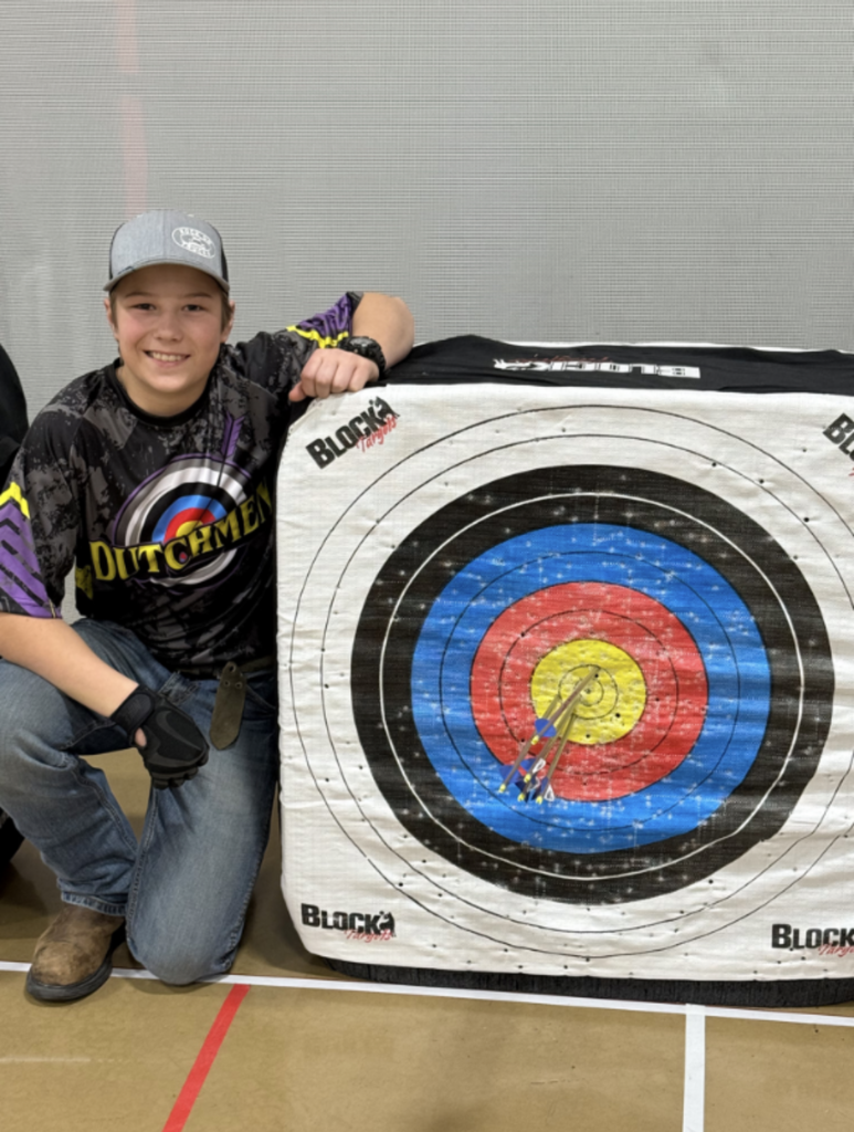 boy with an archery jersey, cap, jeans and boots posed by an archery target with a perfect score