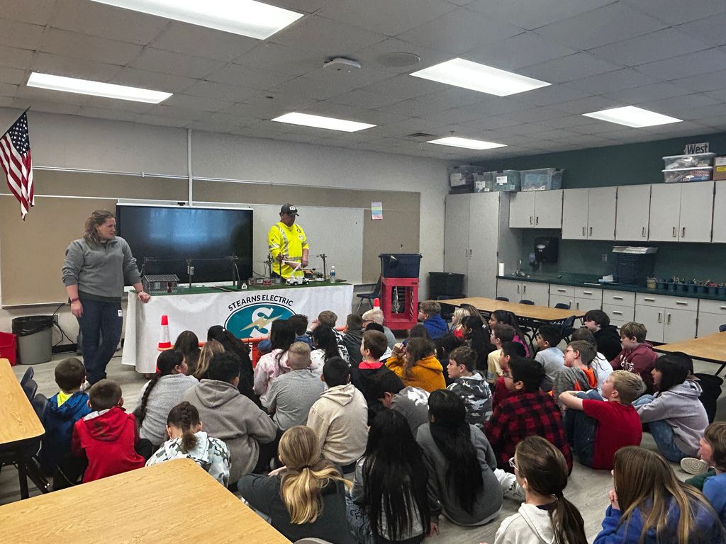 Children sitting on floor facing front with two adults in front . One woman wearing grey sweatshert and one man wearing yellow safety shirt.