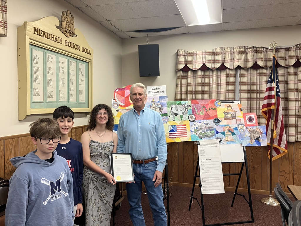 A man and three children stand behind an easel, presenting an award. A flag and a bulletin board are in the background.