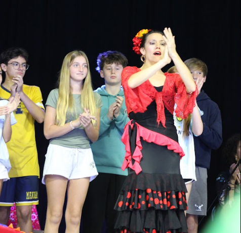 Students standing on stage, one with hands raised and flower in hair, others clapping.