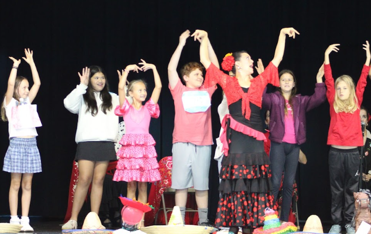 Kids on stage raising hands. One girl in red skirt and hat. Two adults, one wearing red jacket.