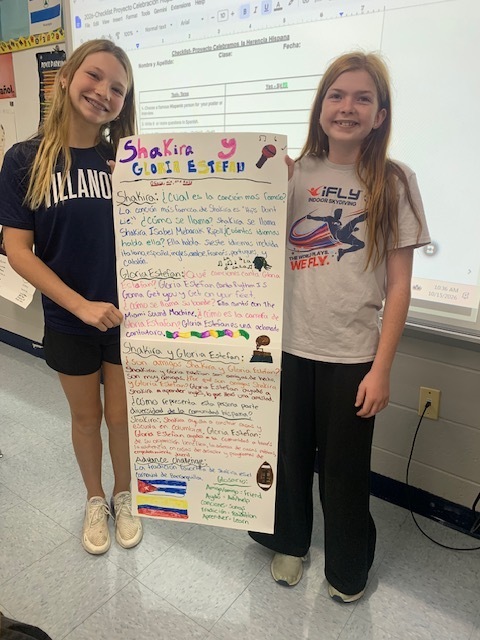 Two girls holding a colorful poster with text, drawings, and pictures in front of a whiteboard.