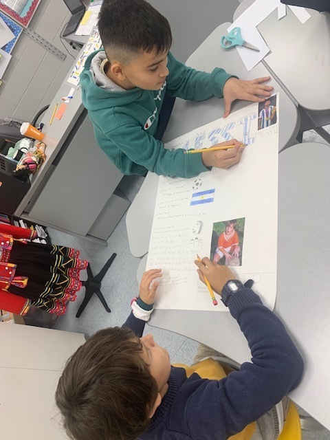 Two children seated at a table, one holding a pen and pointing at a poster.