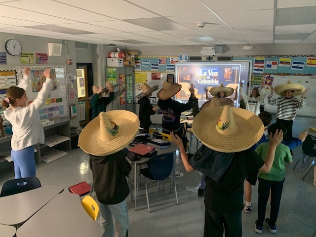People in hats standing in a classroom with desks and chairs. One woman stands in front.