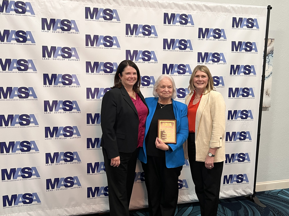 Three women pose before an MASA backdrop; the center woman holds an award plaque while the others smile beside her.