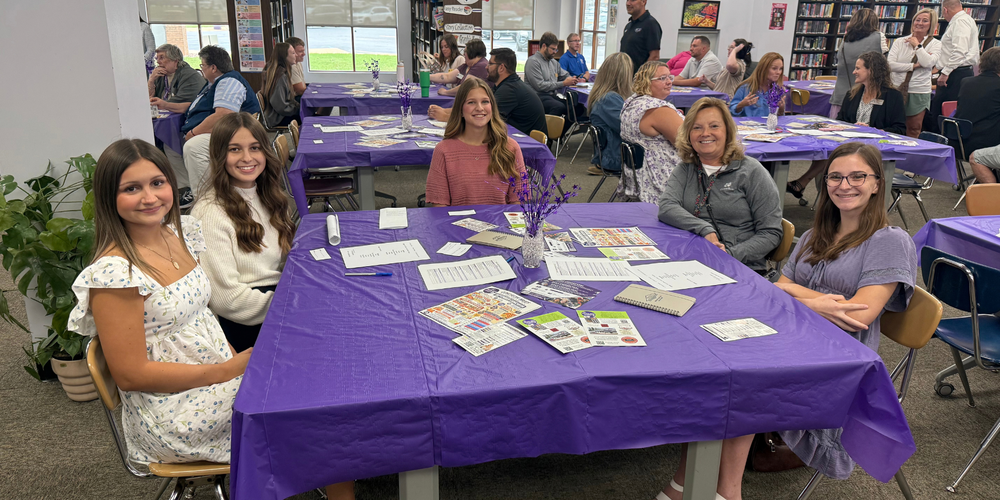 Students and adults gathered at a table with a purple table cover.