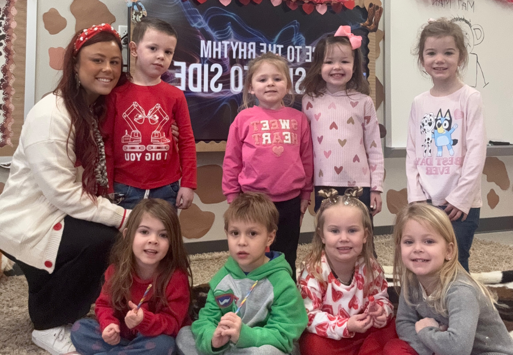 Class photo next to teacher wearing Valentines Day colors, pink and red.