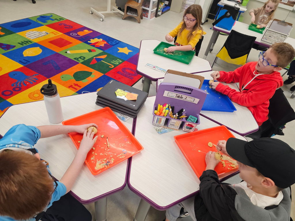 Students knead dough on colorful trays at grouped desks in a bright classroom with supplies and alphabet rug visible.