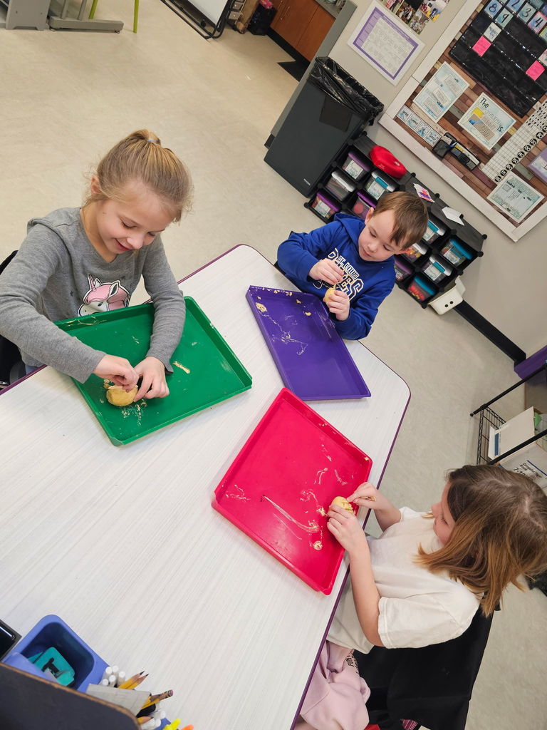 Students knead dough on colorful trays at grouped desks in a bright classroom with supplies.