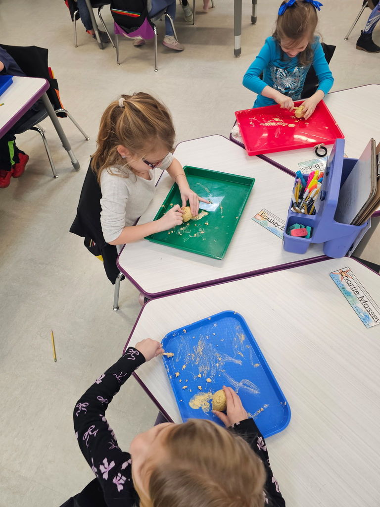 Students knead dough on colorful trays at grouped desks in a bright classroom with supplies.