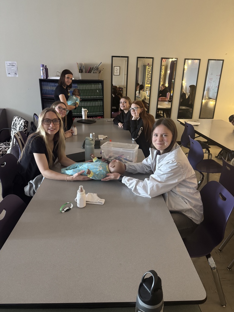 Students gathered around a classroom table practice baby care using dolls, bottles, and blankets during a lesson.