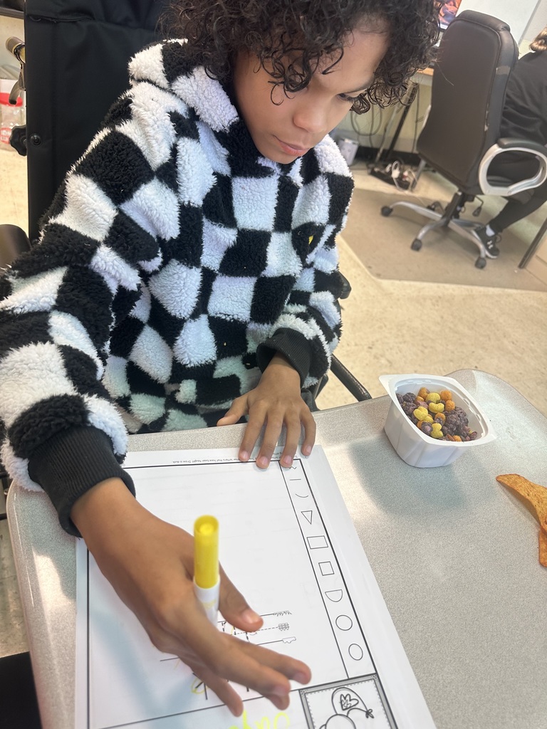 Student in a black-and-white checkered hoodie focuses on a worksheet, using a marker while cereal and a snack sit on the desk nearby.
