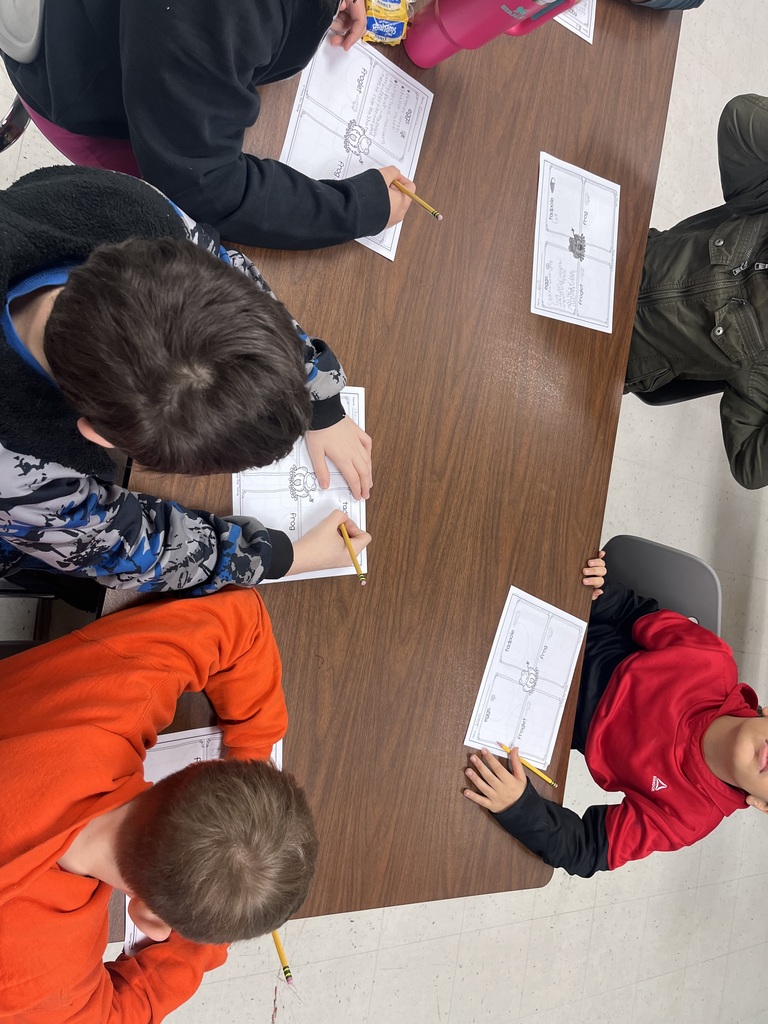 Overhead view of elementary students sitting around a table, writing on worksheets with pencils during a classroom activity.