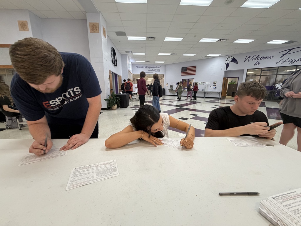Three students filling out forms on a white table inside a school lobby.