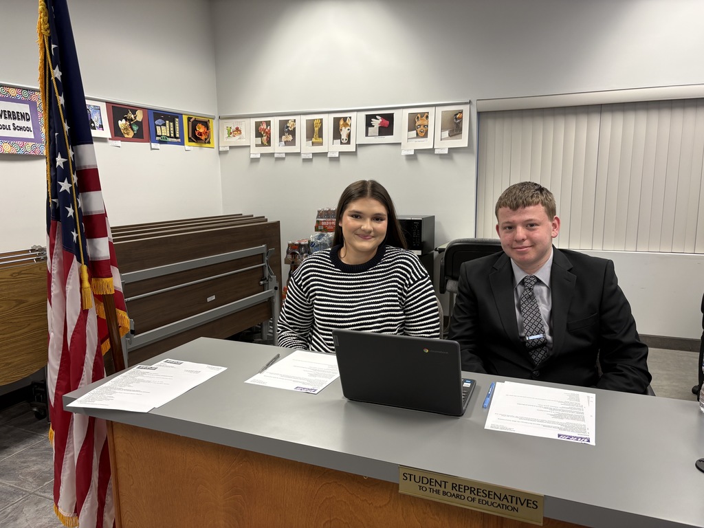 Two students sit at a desk with a laptop and papers, smiling in a school board room; an American flag stands beside them.