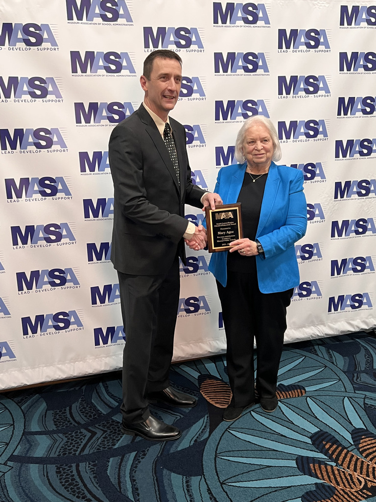 A man shakes hands with an older woman holding an award plaque, both smiling in front of an MASA backdrop.
