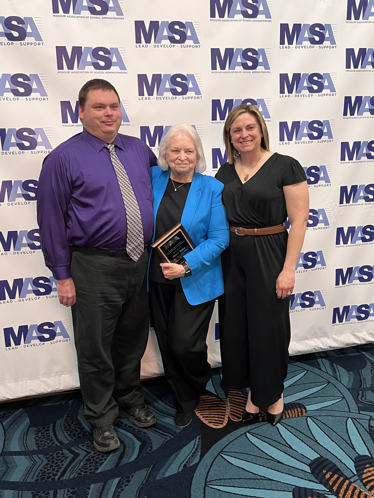 Three people pose in front of an MASA backdrop; an older woman at center holds an award plaque, flanked by two smiling colleagues.