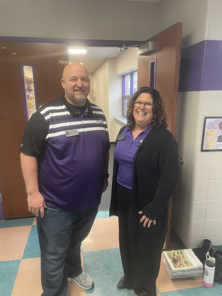 Two smiling board members wearing purple shirts standing in a hallway.