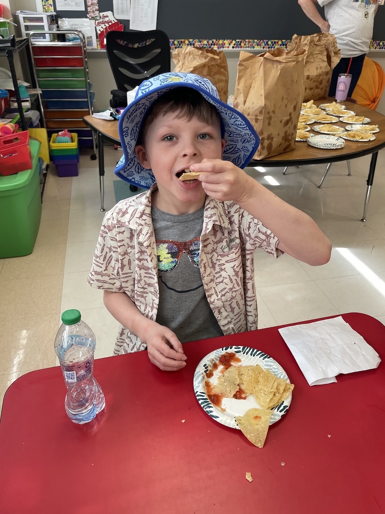 A young student in Hawaiian-themed clothing eats a snack in a classroom.