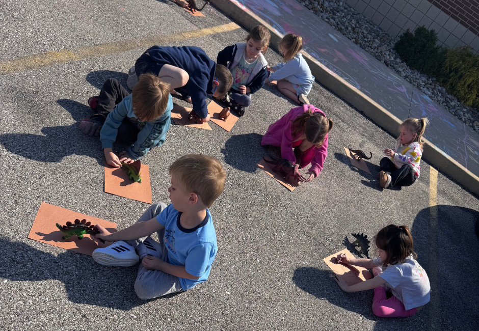 Students playing with dinosaur toys outside with chalk in the background.
