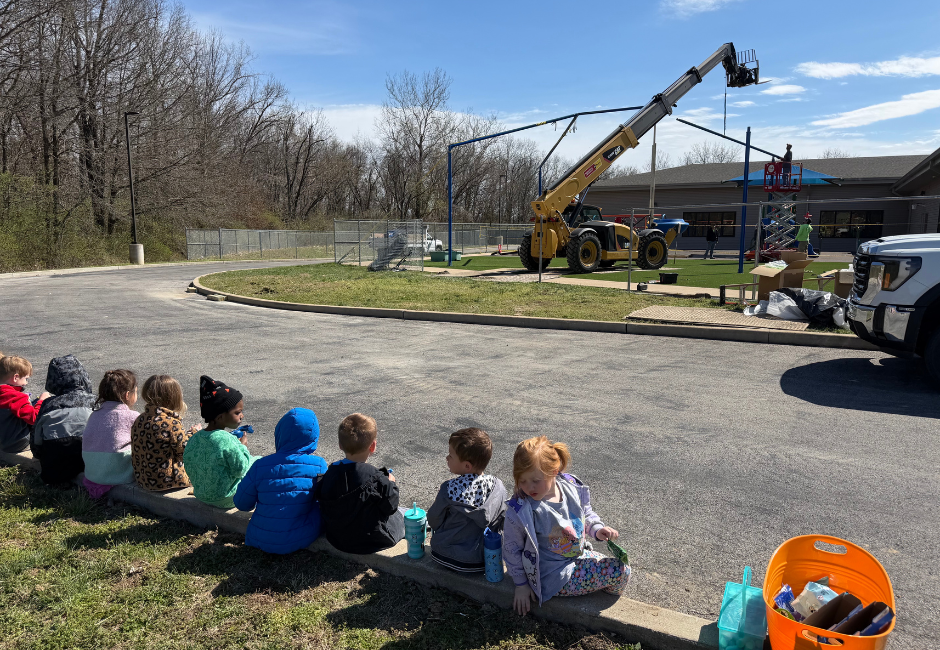 Students sitting on a curb outside watching a yellow machine.