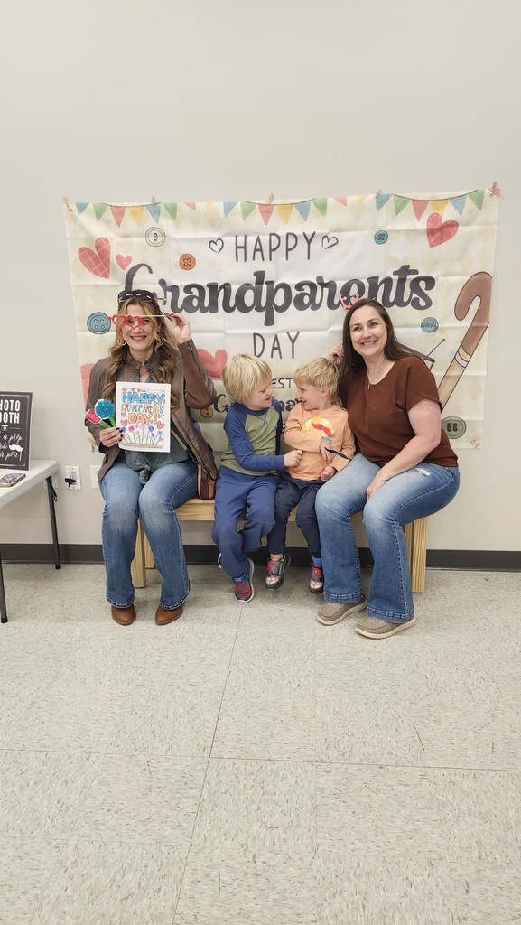 Two students and two adults sitting in front of a "Happy Grandparents Day," banner.