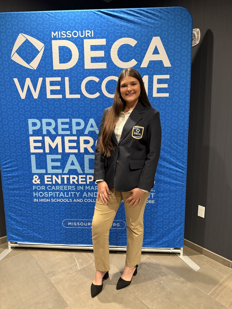 Female student standing in front of a blue banner wearing a black blazer and a tag labeled "DECA."