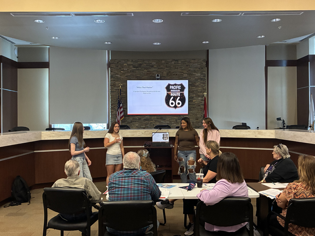Four students standing before a group of adults presenting a slide deck on a big screen.